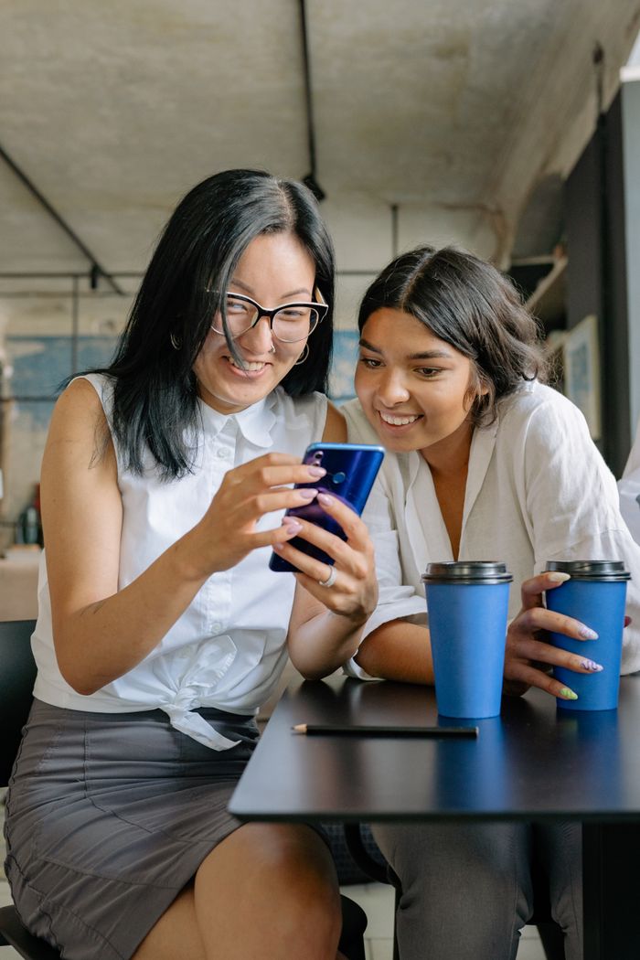 Two women laughing and looking at a smartphone during a coffee break in an office setting.